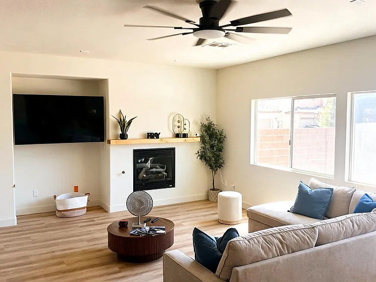 Spacious living room with a fireplace, mounted TV nook, ceiling fan, and large windows overlooking the backyard.