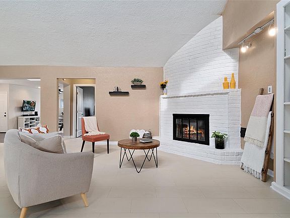 A sky-light and high ceiling soar above the living space that features a white-brick gas fireplace, built-in shelving, and neutral-colored tiles. Natural light exudes through the room.