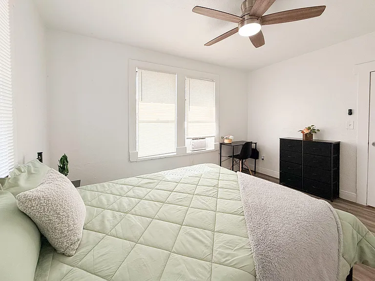 Furnished bedroom with a ceiling fan, two windows with blinds, a work desk, and dresser storage.