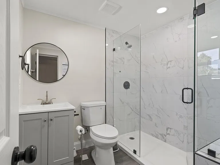 Updated bathroom with a single-sink vanity, round mirror, and walk-in shower with glass enclosure and tiled surround.