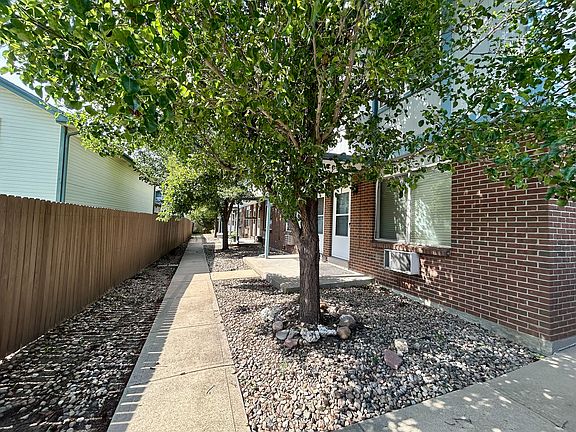 Charming walkway lined with lush green trees leading to a brick apartment building, offering a serene and inviting residential atmosphere.