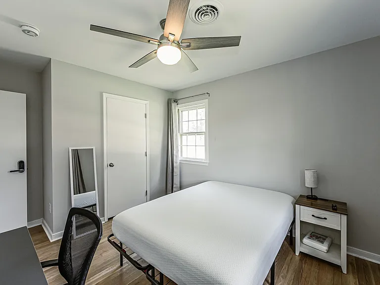 Private bedroom with ceiling fan, window with curtains, and space for a bed and work setup, plus a nightstand.
