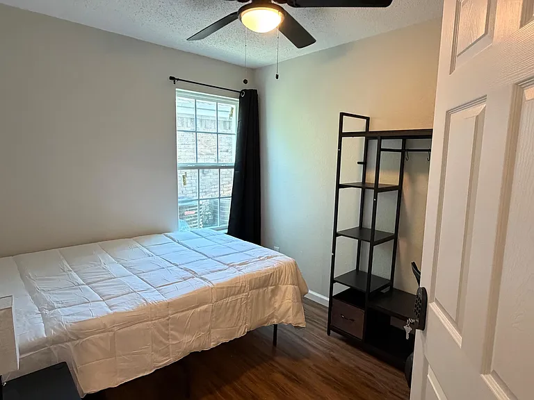 Private furnished bedroom with ceiling fan, wood-look floors, window with curtains, and open shelving for easy storage.