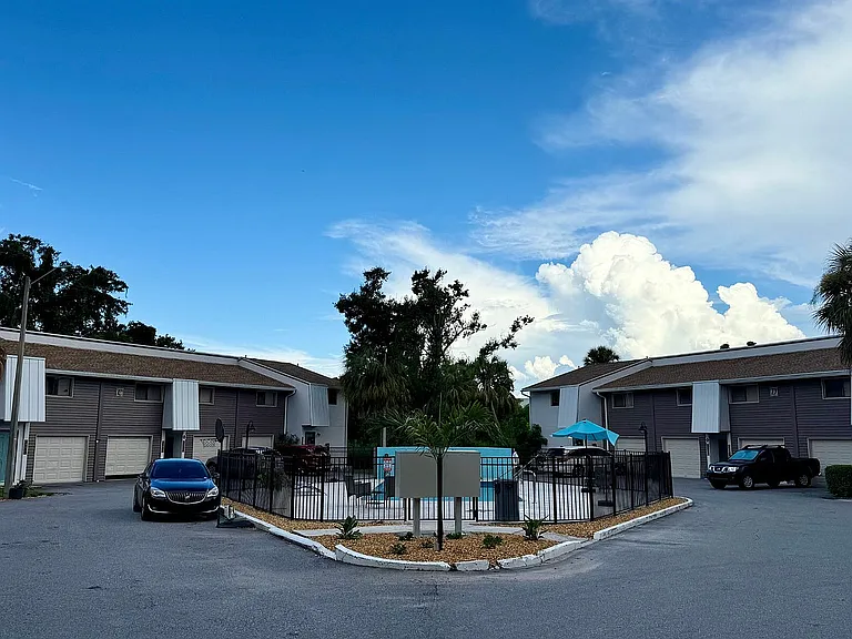 Charming apartment complex with stylish gray exteriors, featuring a landscaped courtyard and inviting pool area under a vibrant blue sky, offering a serene and welcoming environment.