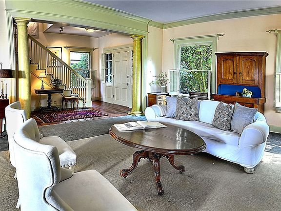 Living room features decorative pillars, crown molding, wood wrapped window with half shutters for privacy, wood burning fireplace with tile surround, decorative columns and classic period details, and carpet flooring.