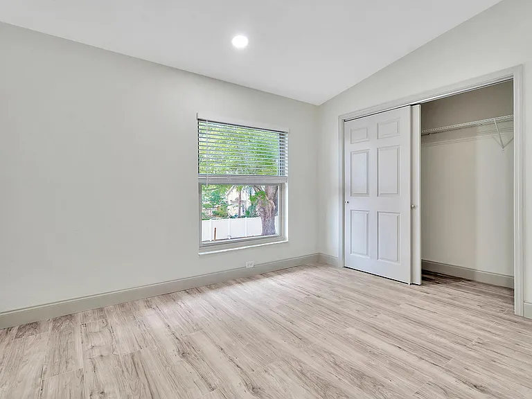Bright, airy bedroom with elegant wood flooring and a large window offering natural light. Features a spacious closet with sliding doors.