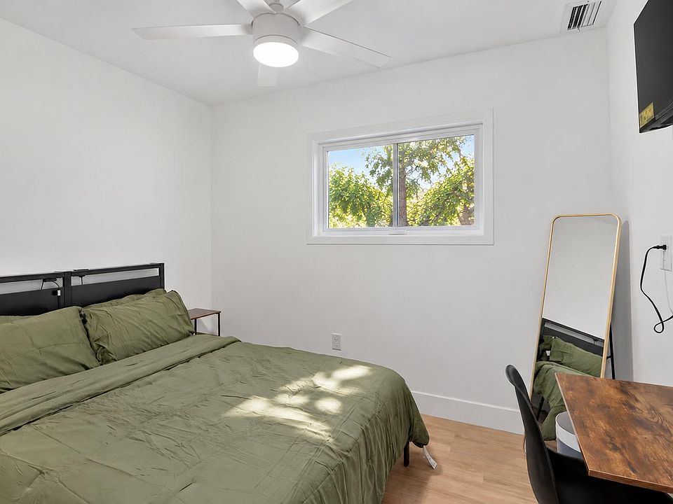 Bedroom offers a comfortable layout with a large window, ceiling fan, wall-mounted TV, and a dedicated desk area.