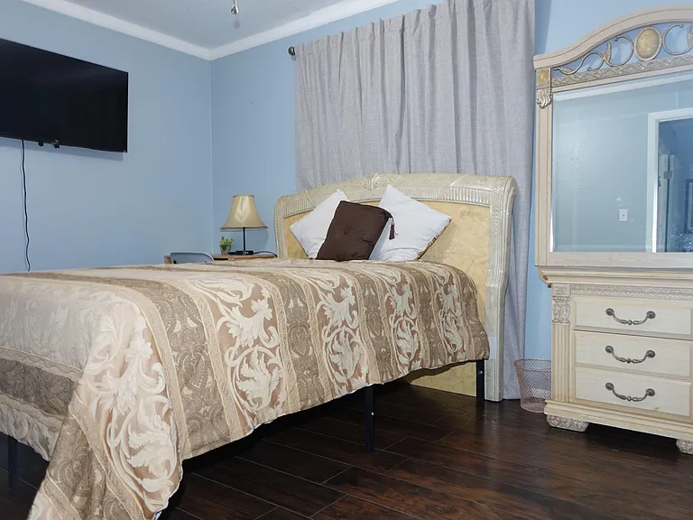Furnished bedroom with a queen bed, nightstand, and dresser/mirror set, plus a wall-mounted TV and window curtains.
