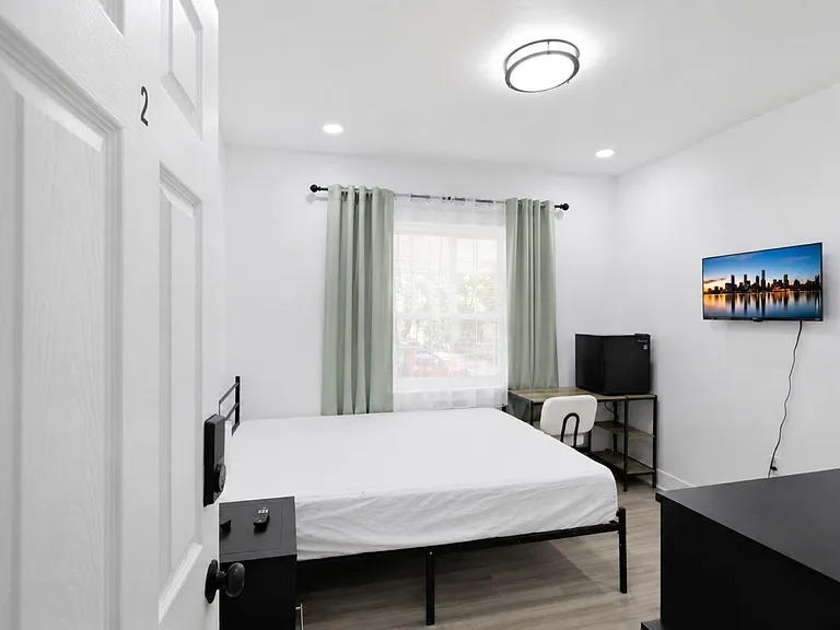 Private bedroom with a full bed, window with curtains, work desk, mini fridge, and wall-mounted TV for easy living.