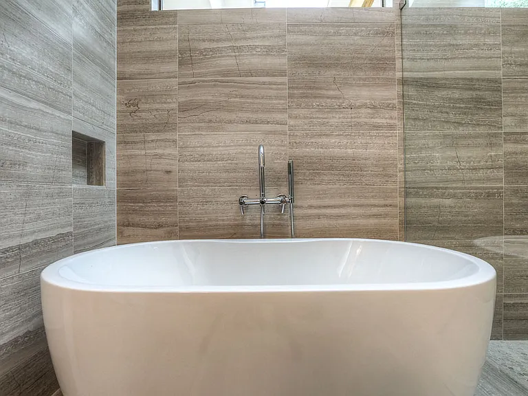 Detail of freestanding soaking tub with chrome floor-mount filler against floor-to-ceiling stone-look tile
