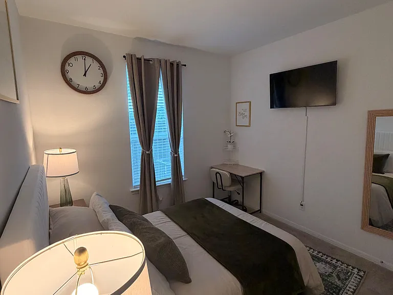 Furnished bedroom with a full bed, nightstand lighting, window with curtains, a compact desk, and wall-mounted TV.