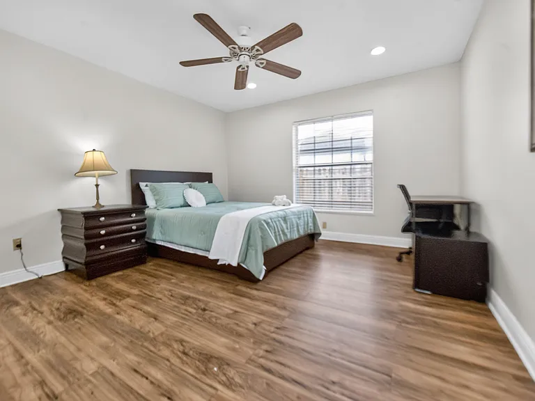 Furnished bedroom with wood-look flooring, ceiling fan, a large window with blinds, and a dedicated desk workspace.