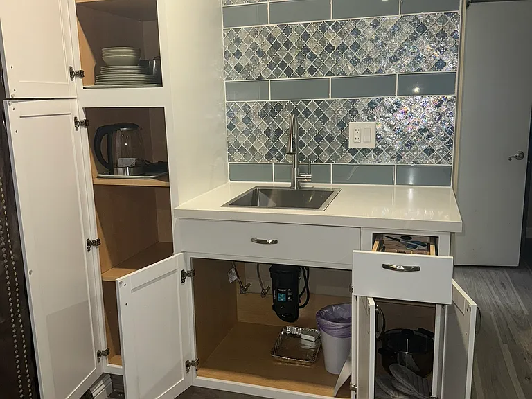 All the cupboards open, there are place settings for 4 people including cups, glasses, plates, silverware, some cooking utensils as well as pots and pans, french press, and drying rack. The sink has a garbage disposal in it