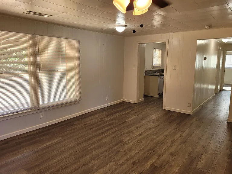 Living room with view into kitchen on left, hallway to bedrooms on right. Newer laminate flooring, ceiling fan and mini blinds.
