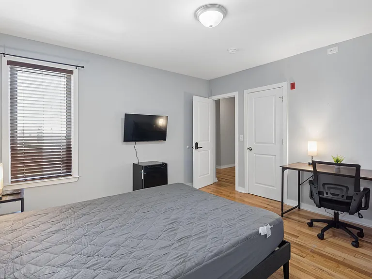 Private bedroom with wood-style floors, large window with blinds, wall-mounted TV, mini fridge, and a work desk area.