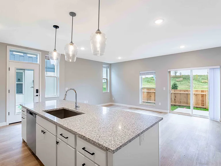 Bright, modern open-concept living space with a sleek granite kitchen island, elegant pendant lighting, and large windows offering a tranquil view of the lush green backyard.
