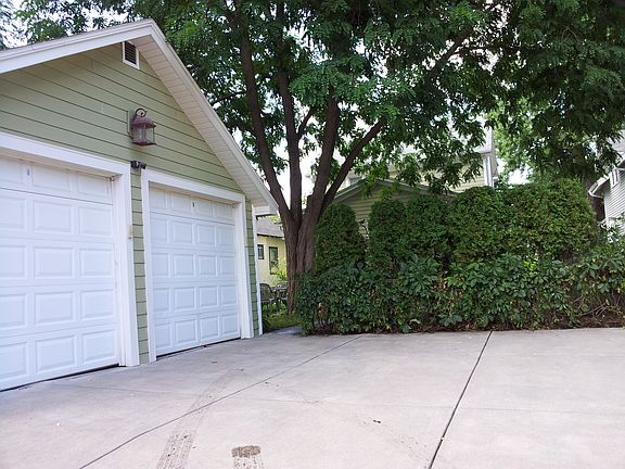 Northwest view/Alley view of 2 garages separated by a wall each with its own garage door fob and key pad this is an old picture there are tall grasses where the arborvitaes are here.