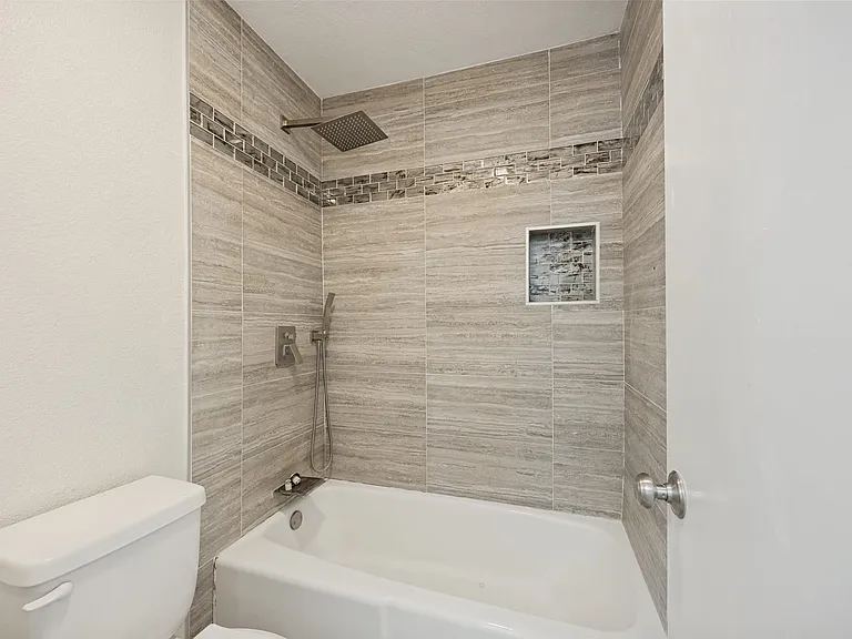 Bathroom featuring a tub/shower combination with large format tiled walls, a decorative mosaic tile accent band, a built-in shower niche, and a rain showerhead with a handheld sprayer