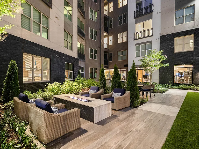 Elegant courtyard in a modern apartment complex featuring cozy seating around a sleek outdoor fire pit, surrounded by lush greenery and well-lit windows.