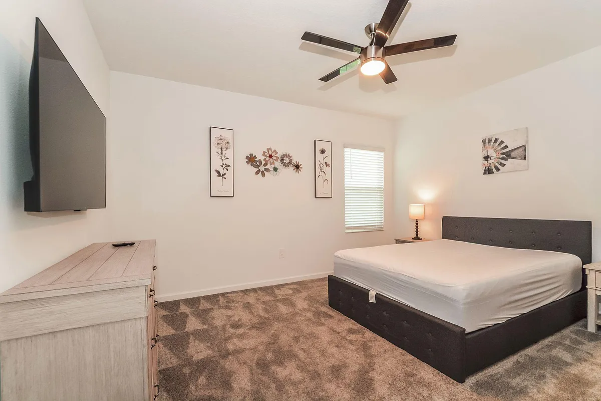Spacious bedroom with a ceiling fan, wall-mounted TV, dresser storage, and a window with blinds for privacy. - Slash Way