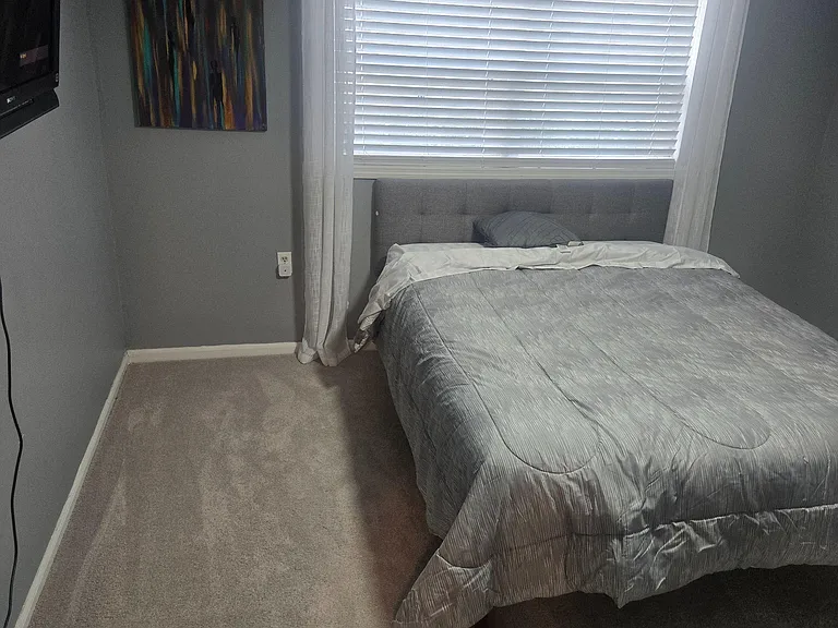 Furnished bedroom with a full-size bed, upholstered headboard, and window with blinds and curtains for privacy.