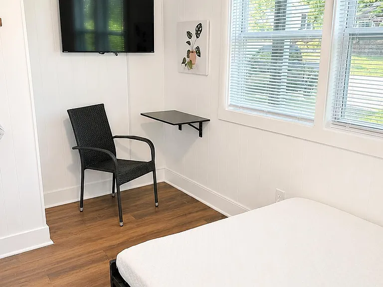 Private bedroom with wood-look flooring, large corner windows with blinds, and a wall-mounted TV plus a small work surface.