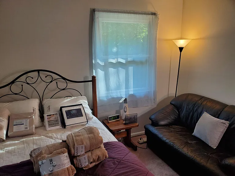 Furnished corner bedroom featuring two windows with curtains, nightstand, and a comfortable seating sofa for relaxing.