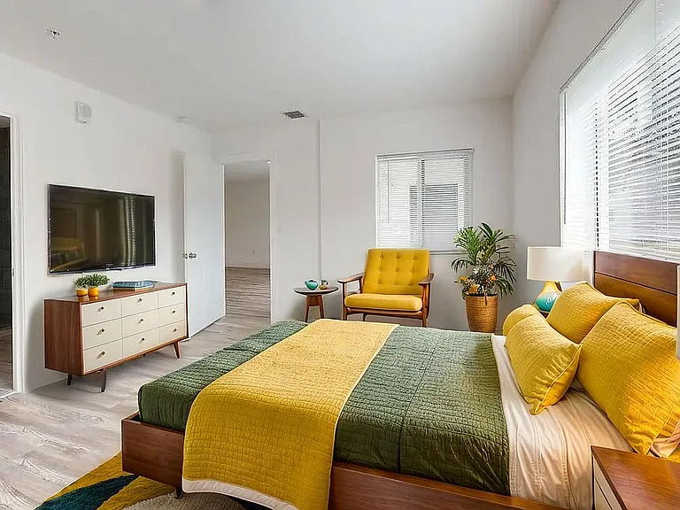 Bright and modern bedroom featuring a stylish bed with vibrant yellow and green bedding, complemented by a cozy armchair and elegant chest of drawers beneath a sleek wall-mounted TV.