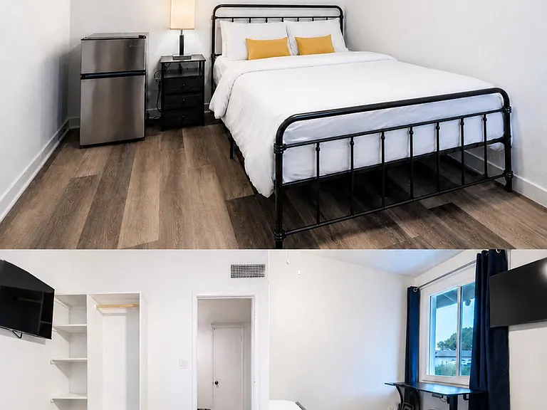 Furnished bedroom with queen bed, mini fridge, desk workspace, open shelving, and wood-look flooring.