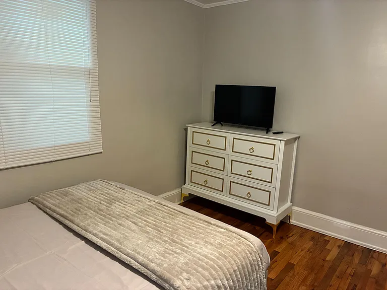 Bedroom featuring a window with blinds, hardwood floors, and a dresser with TVideal for relaxing and added storage.