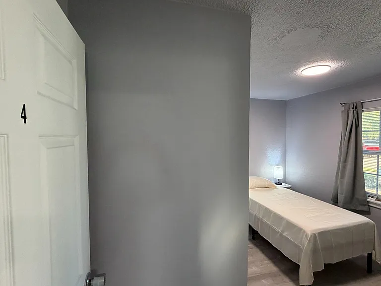 Private bedroom with a furnished bed, bedside table and lamp, plus a window with blinds and curtains for light control.