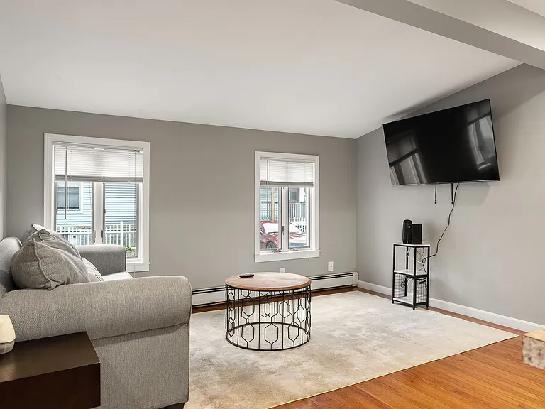 Living Area, Wall-Mounted TV Setup, Hardwood Floors, Area Rug, Natural Light, Neutral Gray Walls, Baseboard Heating.