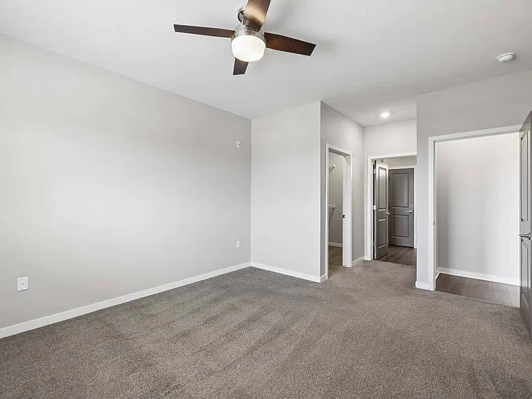 Spacious apartment bedroom with carpeted flooring, a ceiling fan, neutral walls, and an open layout with hallway access and a sliding closet door.