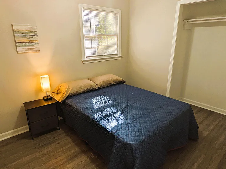 Furnished bedroom with a full-size bed, nightstand and lamp, plus a window with blinds and an open closet for storage.