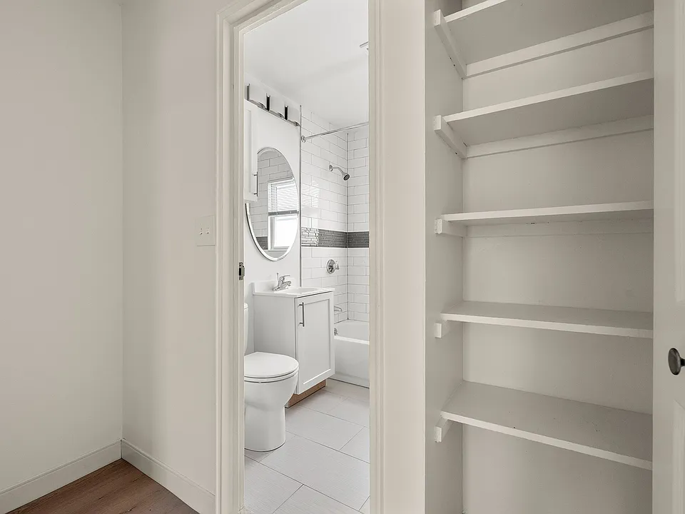Modern bathroom with sleek white tiles, a stylish round mirror, and an adjacent open shelving unit for ample storage.