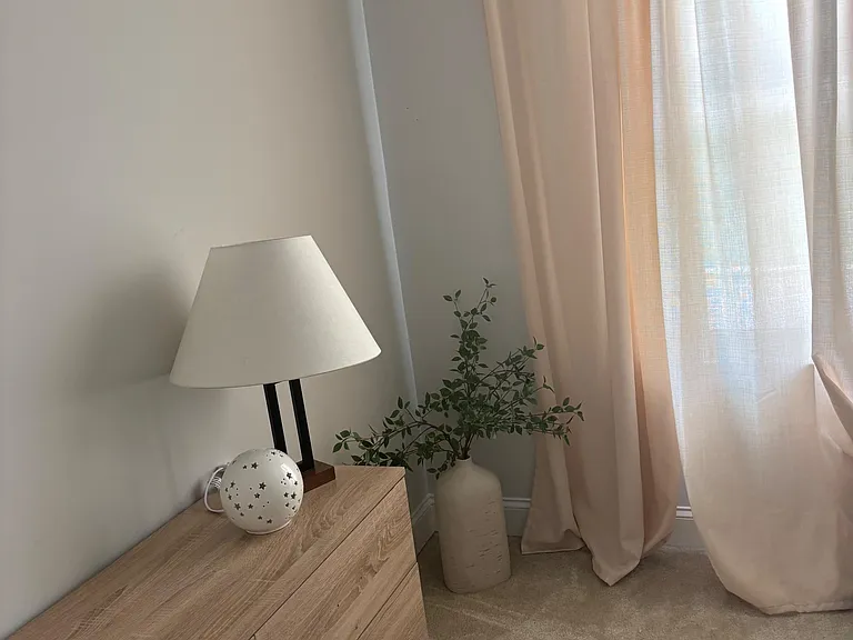 Carpeted bedroom corner with a dresser, bedside lighting, and a large window with curtains for natural light.