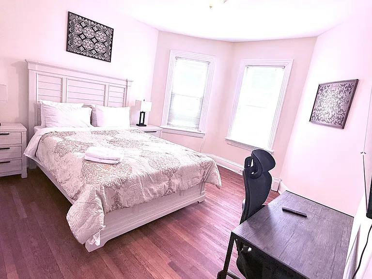 Furnished bedroom with a full bed, nightstands, and a dedicated desk area, plus two windows and hardwood floors.