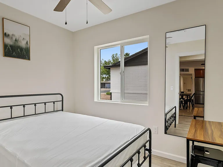 Private bedroom with a full-size bed, large window, ceiling fan, and a compact desk area with mirror for daily prep.
