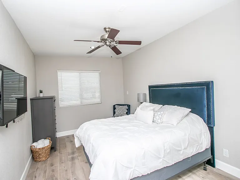 Detached casita bedroom at, Tempe rental home. Features a queen bed with a tufted navy velvet headboard, white bedding, a wall mounted flatscreen TV, a tall dresser, wood-plank flooring, and ceiling fan. Comfortable bedroom suite for a top Tempe, AZ stay.
