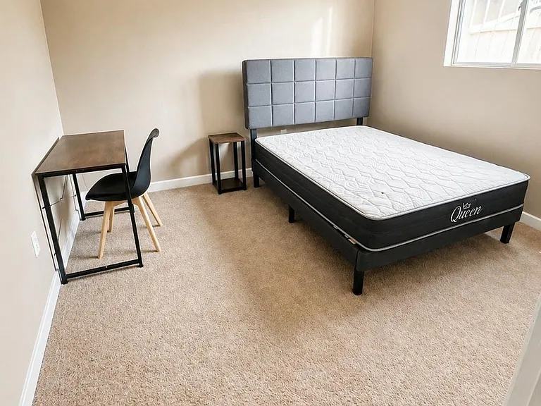 Furnished bedroom with a queen bed, nightstand, and dedicated desk area, plus a window and soft carpet underfoot.