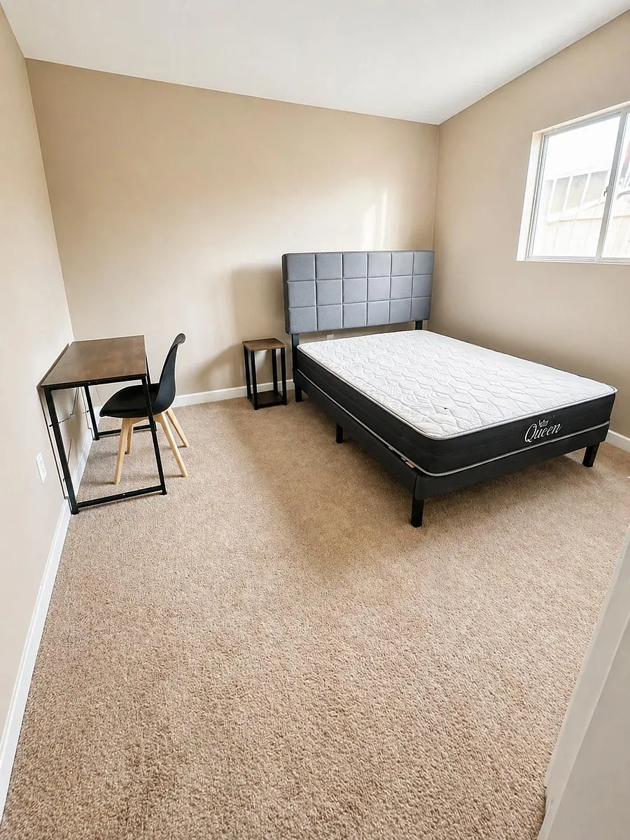 Furnished bedroom with a queen bed, nightstand, and dedicated desk area, plus a window and soft carpet underfoot. - Frederick Ave