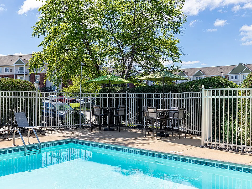 Inviting outdoor pool area with lounge chairs and shaded tables, surrounded by lush greenery and modern apartments.