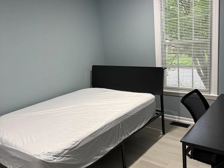 Private bedroom with a furnished bed, workspace desk, and a window with blinds for natural light and privacy.