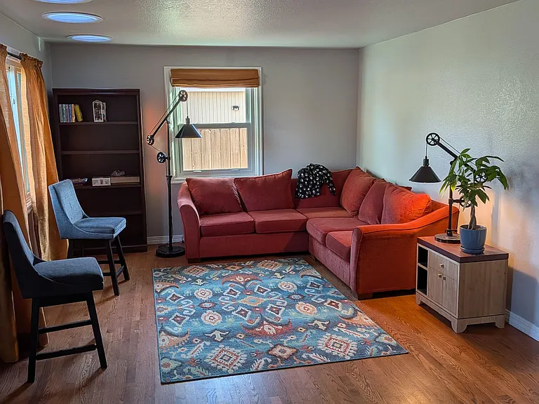 Living room with sectional seating, accent chairs, bookcase storage, and hardwood-style floors with two windows.