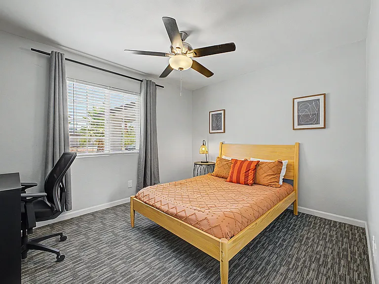 Furnished bedroom with a full bed, work desk, and ceiling fan, plus a large window with blinds and curtains.
