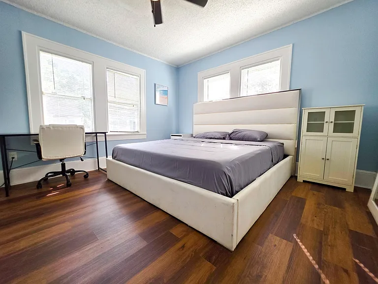 Spacious bedroom with hardwood floors, three windows, a queen bed, and a dedicated desk area for work or study.