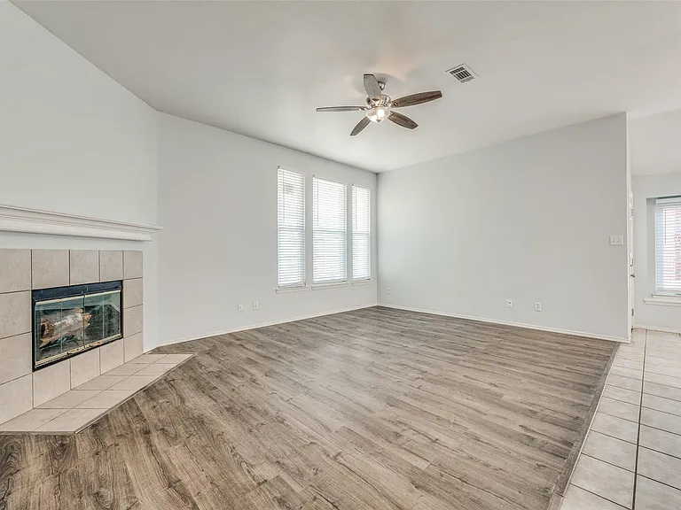 Spacious living area featuring wood-finish flooring, a tiled fireplace with mantel, multiple windows with blinds, and a ceiling fan with integrated lighting