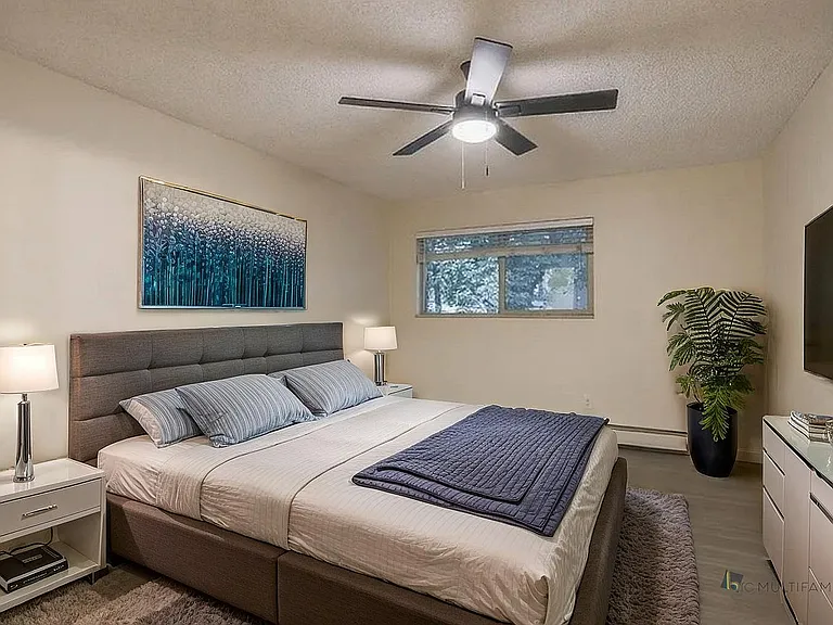 Inviting bedroom with plush bedding, stylish decor, and natural light from a large window; features sleek furnishings and a calming plant for a serene atmosphere.