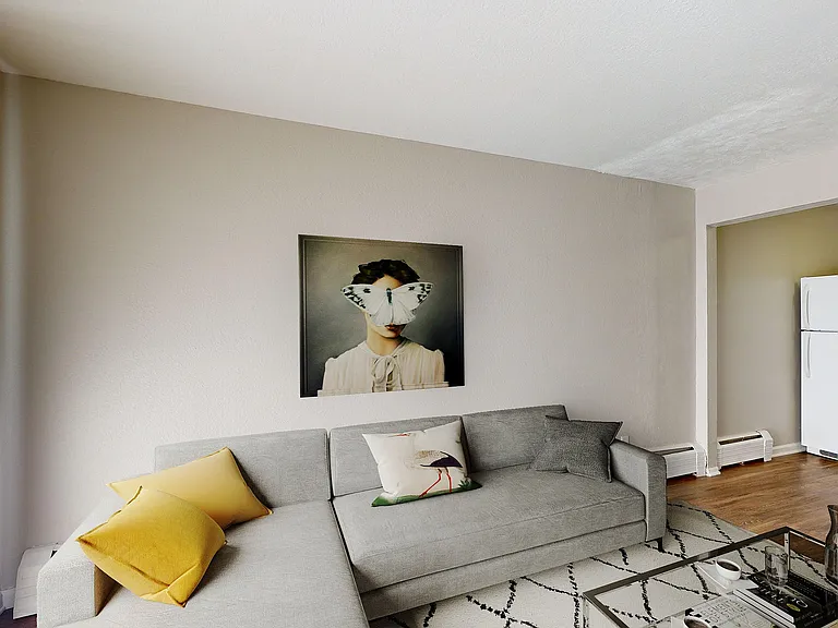 Modern living room with a stylish gray sofa adorned with vibrant yellow and patterned cushions, contemporary art on the wall, and an inviting atmosphere near the kitchen area with natural light streaming through vertical blinds.
