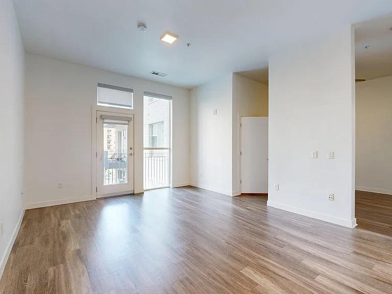 A bright, empty living room with wood-style flooring, white walls, and a glass door leading to a balcony.